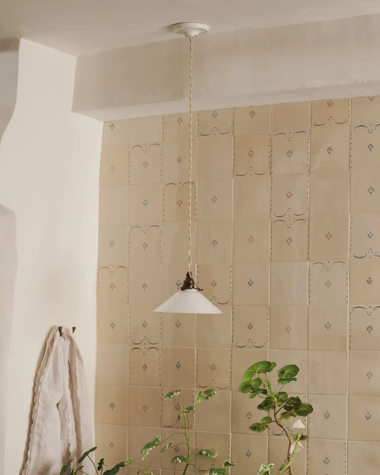 A small pendant light with translucent milk lamp shade in front of a wall filled with ditsy delft tiles pattern, with a console table with potted plants.