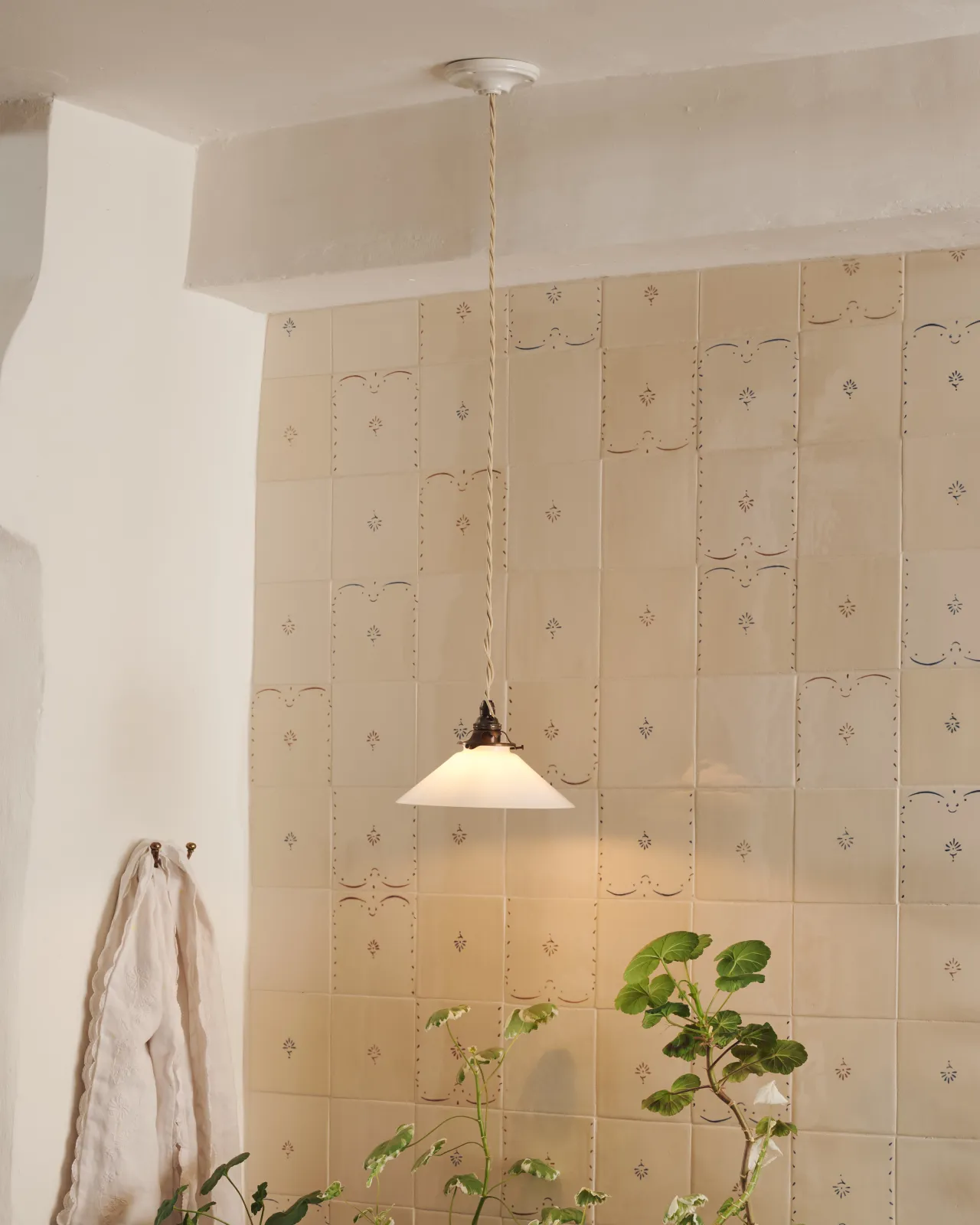 A small pendant light with translucent milk lamp shade in front of a wall filled with ditsy delft tiles pattern, with a console table with potted plants.