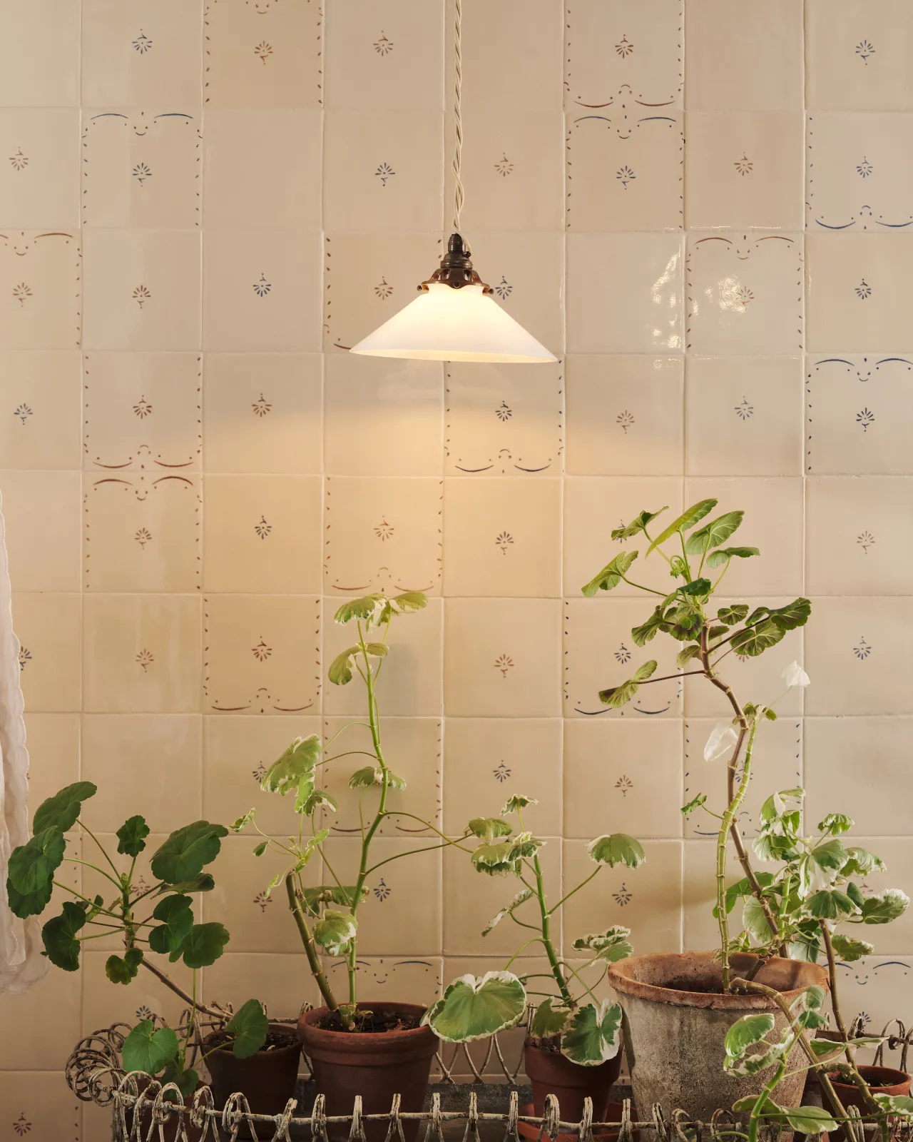 A small pendant light with translucent milk lamp shade in front of a wall filled with ditsy delft tiles pattern, with a console table with potted plants.