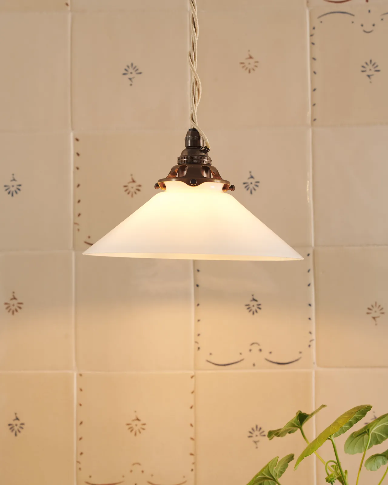 A small pendant light with translucent milk lamp shade in front of a wall filled with ditsy delft tiles pattern, with a console table with potted plants.