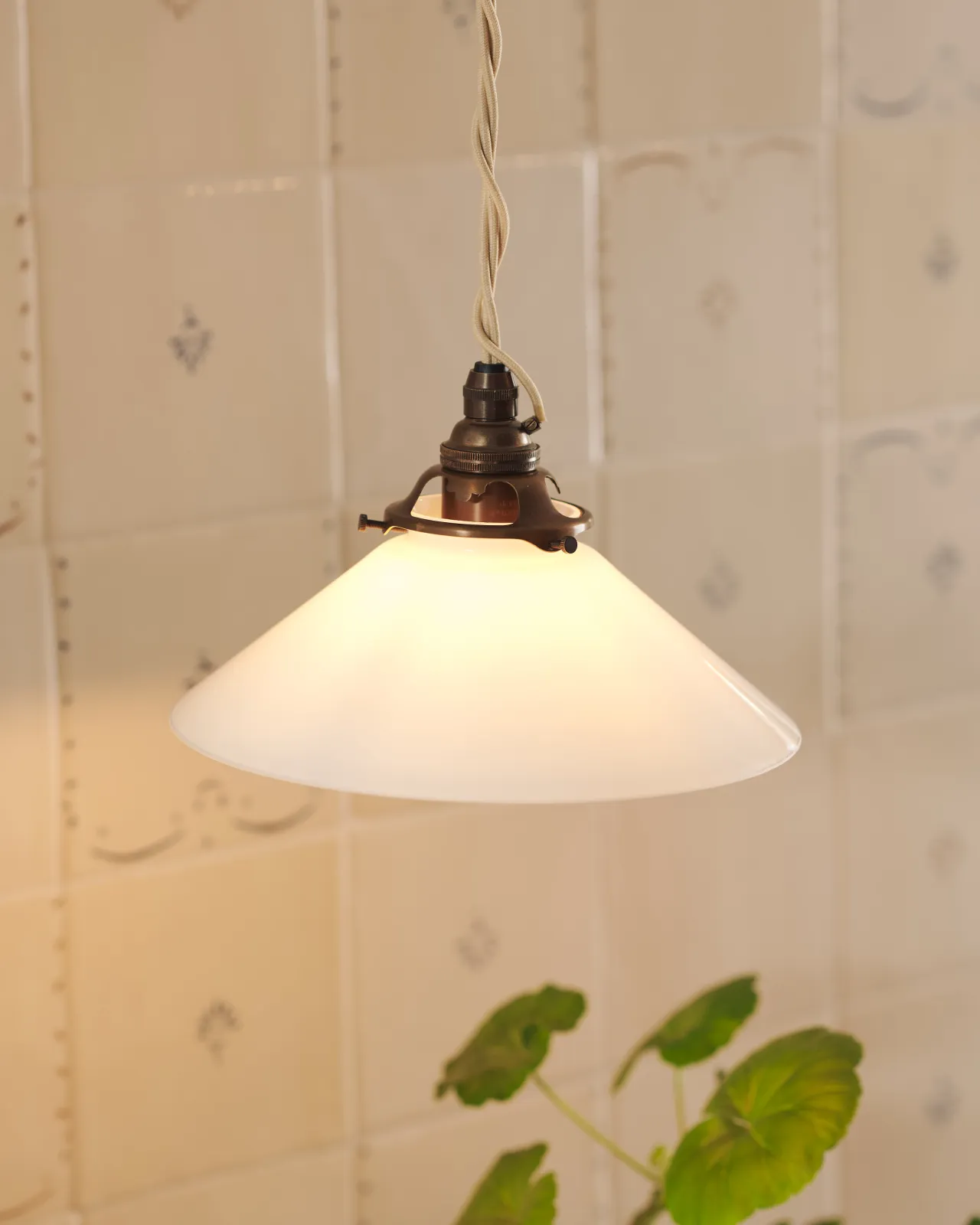 A small pendant light with translucent milk lamp shade in front of a wall filled with ditsy delft tiles pattern, with a console table with potted plants.