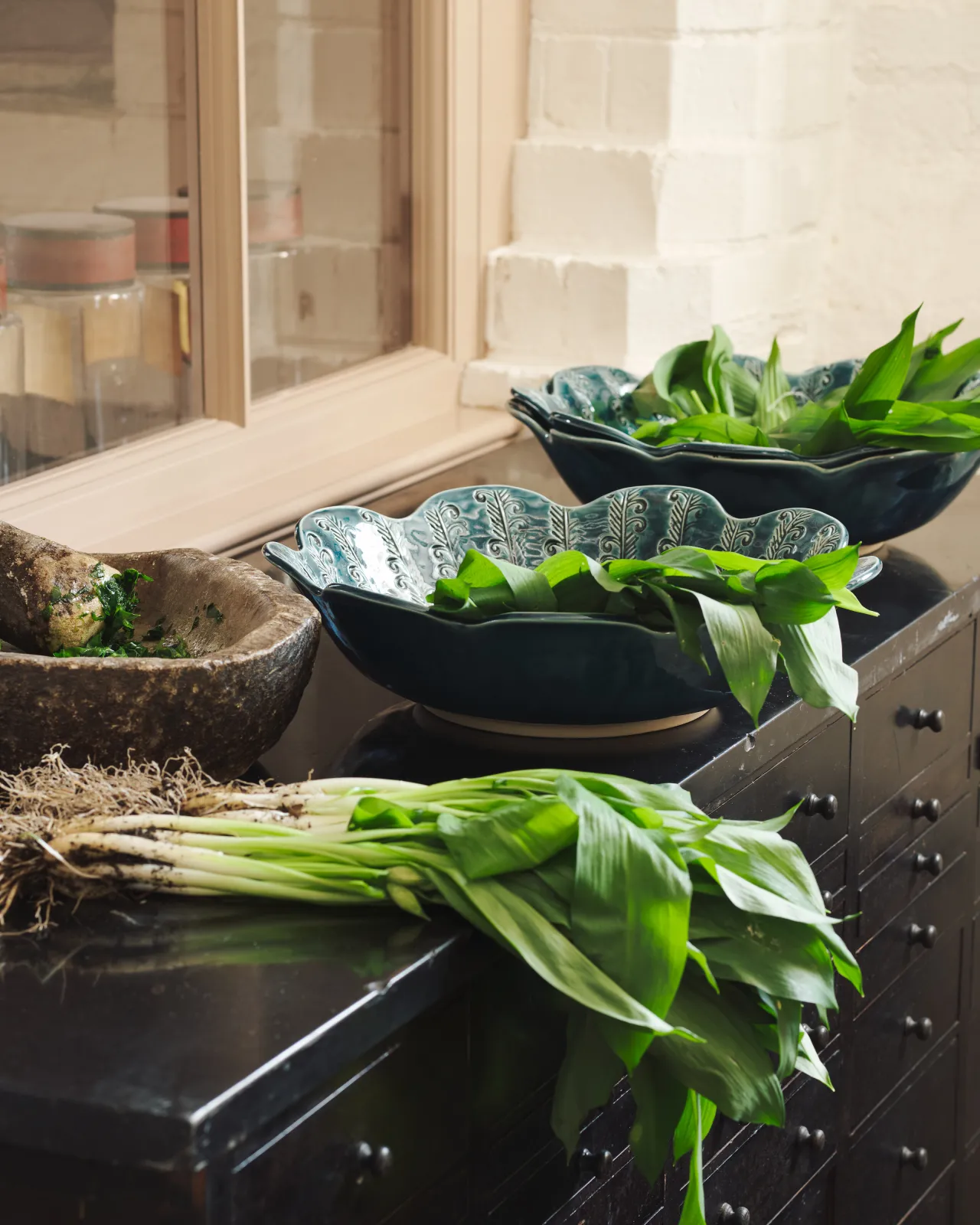 Blue glazed petal-shaped ceramic bowl with embossed pattern, styled with fresh wild garlic on a dark surface.