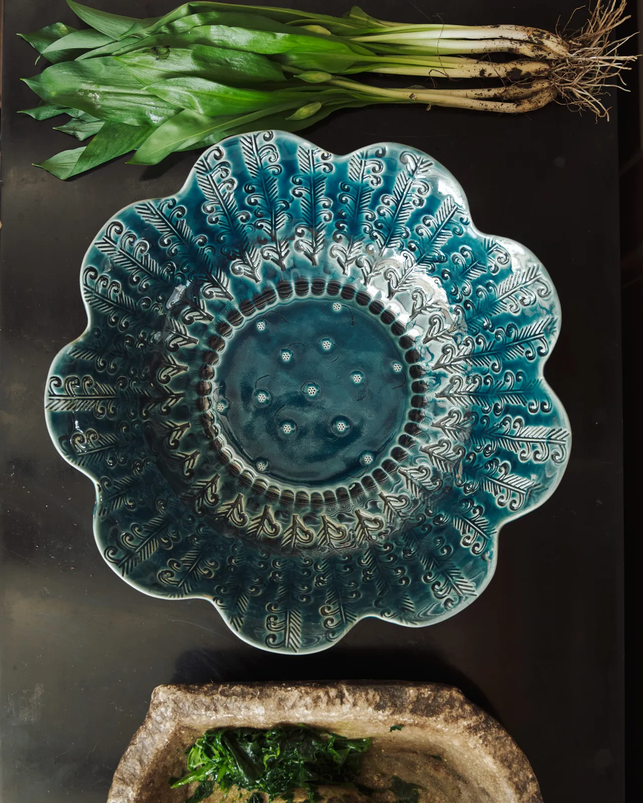 Blue glazed petal-shaped ceramic bowl with embossed pattern, styled with fresh wild garlic on a dark surface.