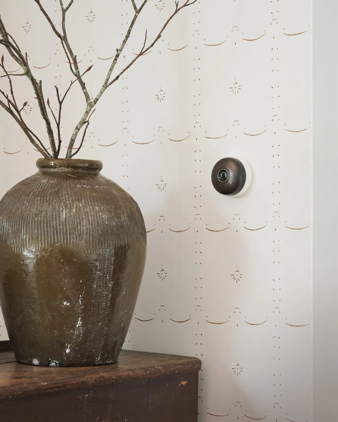 An Oxidised Bronze deVOL Dolly Toggle Switch with a white ceramic base, fitted to a wall of deVOL's Sepia Ditsy Delft wallpaper.