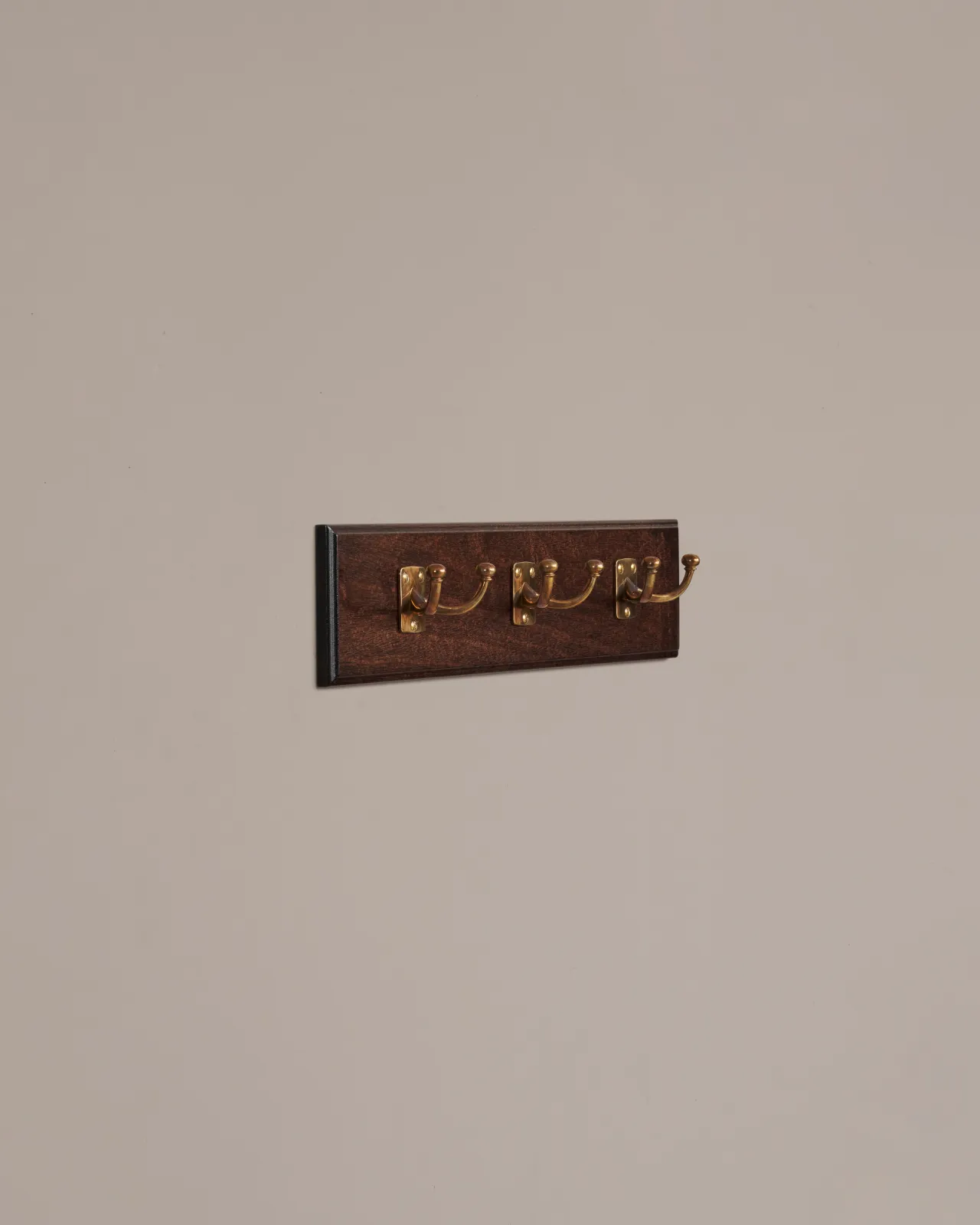 An oblique view of three brass Twin Edwardian Coat Hooks on a Mahogany Finish Backboard on a plain grey wall.