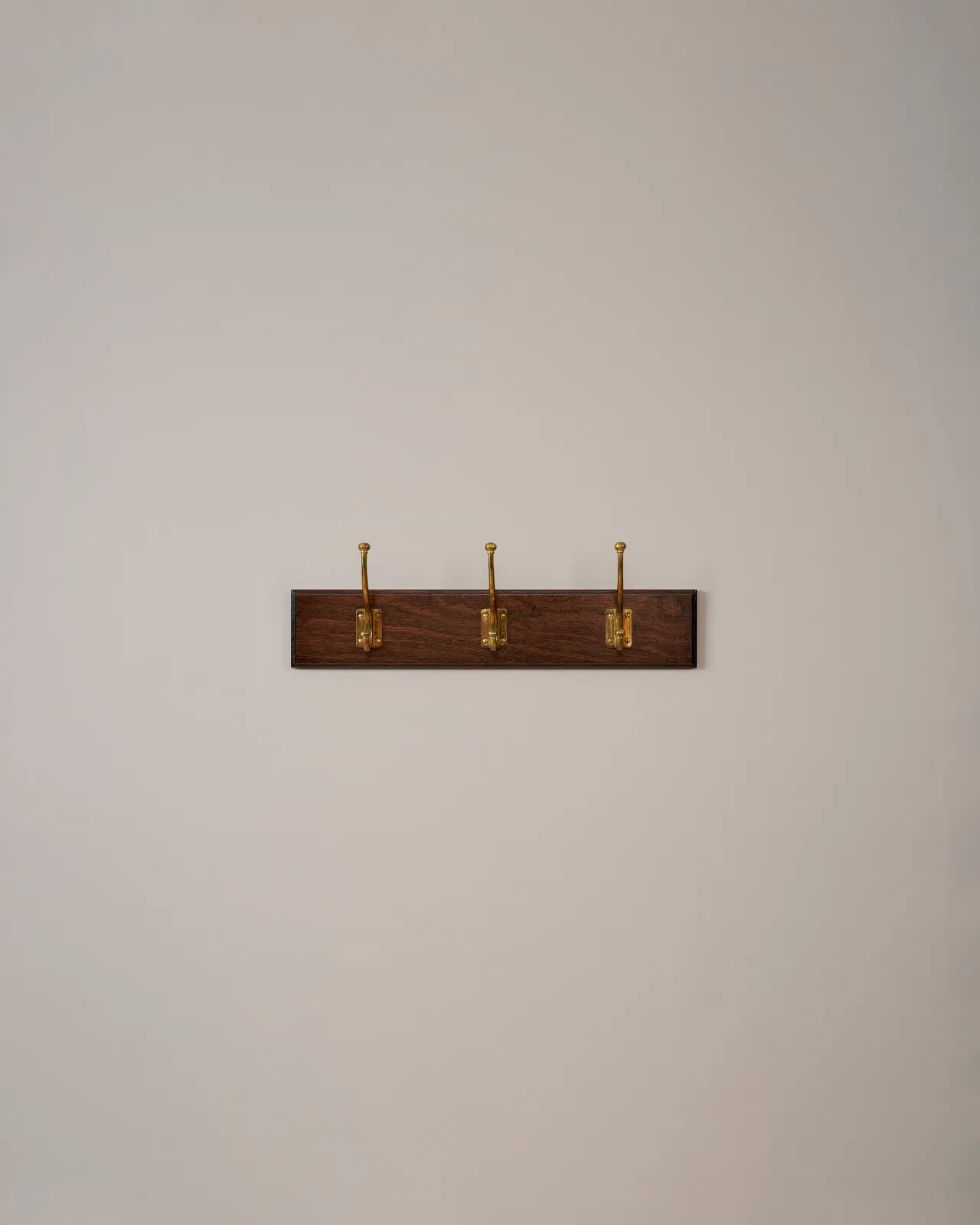 Three brass Edwardian Coat Hooks on a Mahogany Finish Backboard on a plain grey wall.