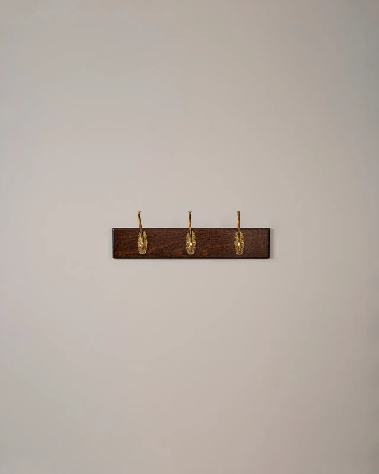 Three brass Petite Parisian Café Hooks on a Mahogany Finish Backboard on a plain grey wall.