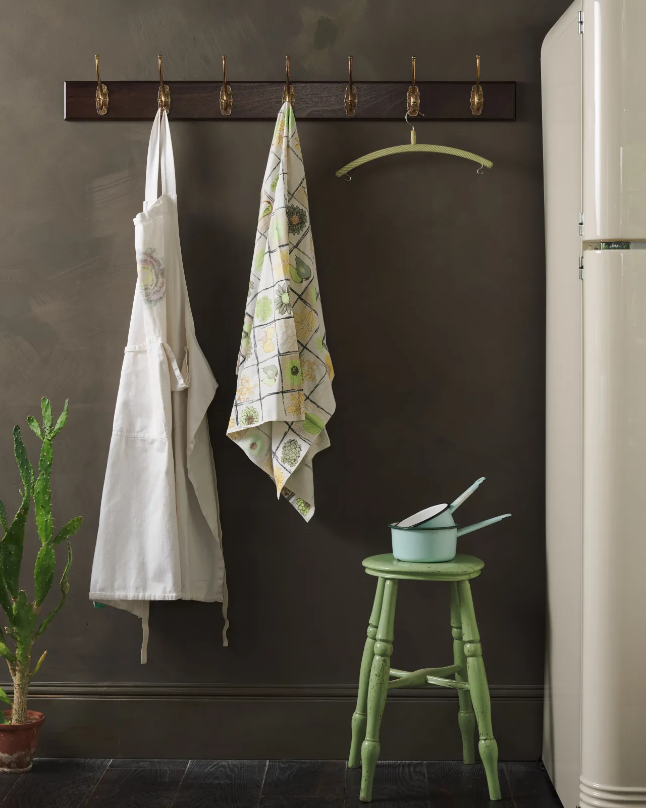 A Mahogany Finish Hook Rack with seven brass Parisian Café Hooks, fitted to a dark wall with two aprons hanging above a rustic green milking stool.