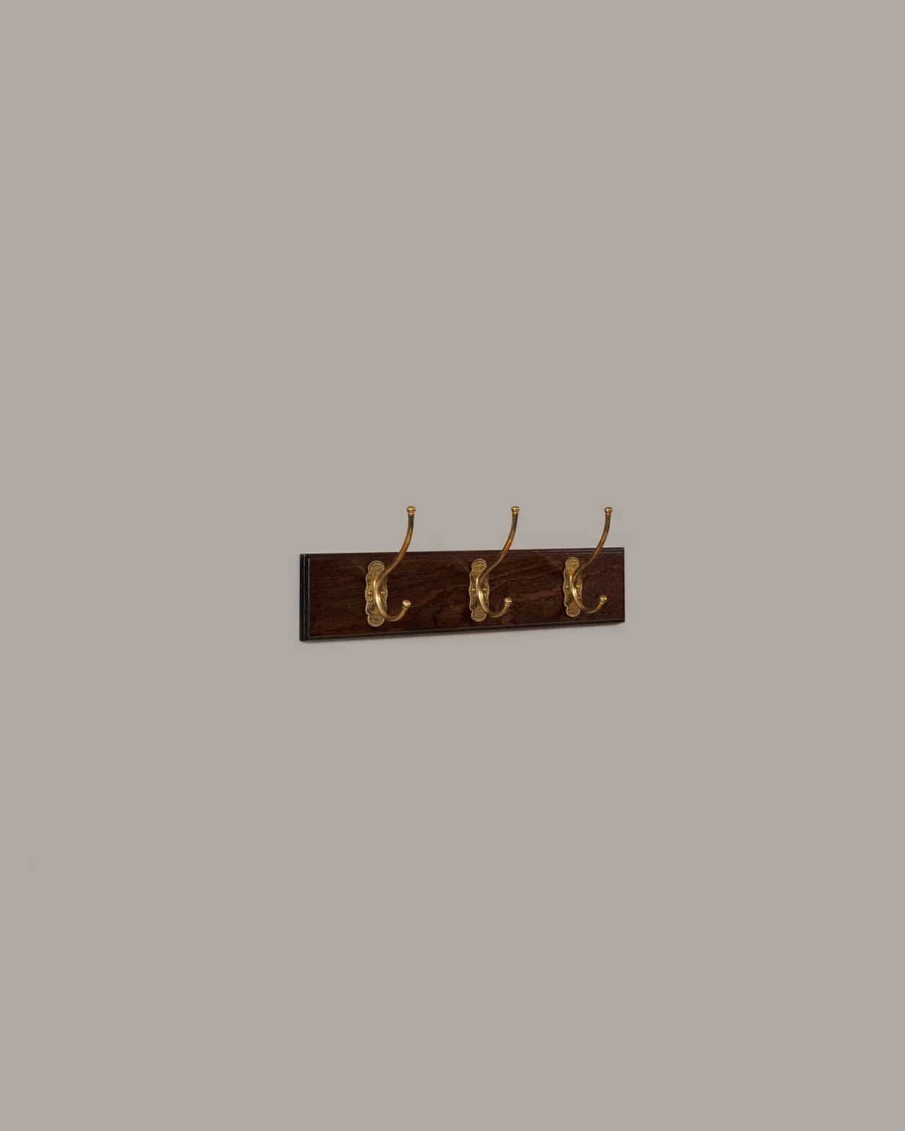 An oblique view of three brass Petite Parisian Coat Hooks on a Mahogany Finish Backboard on a plain grey wall.