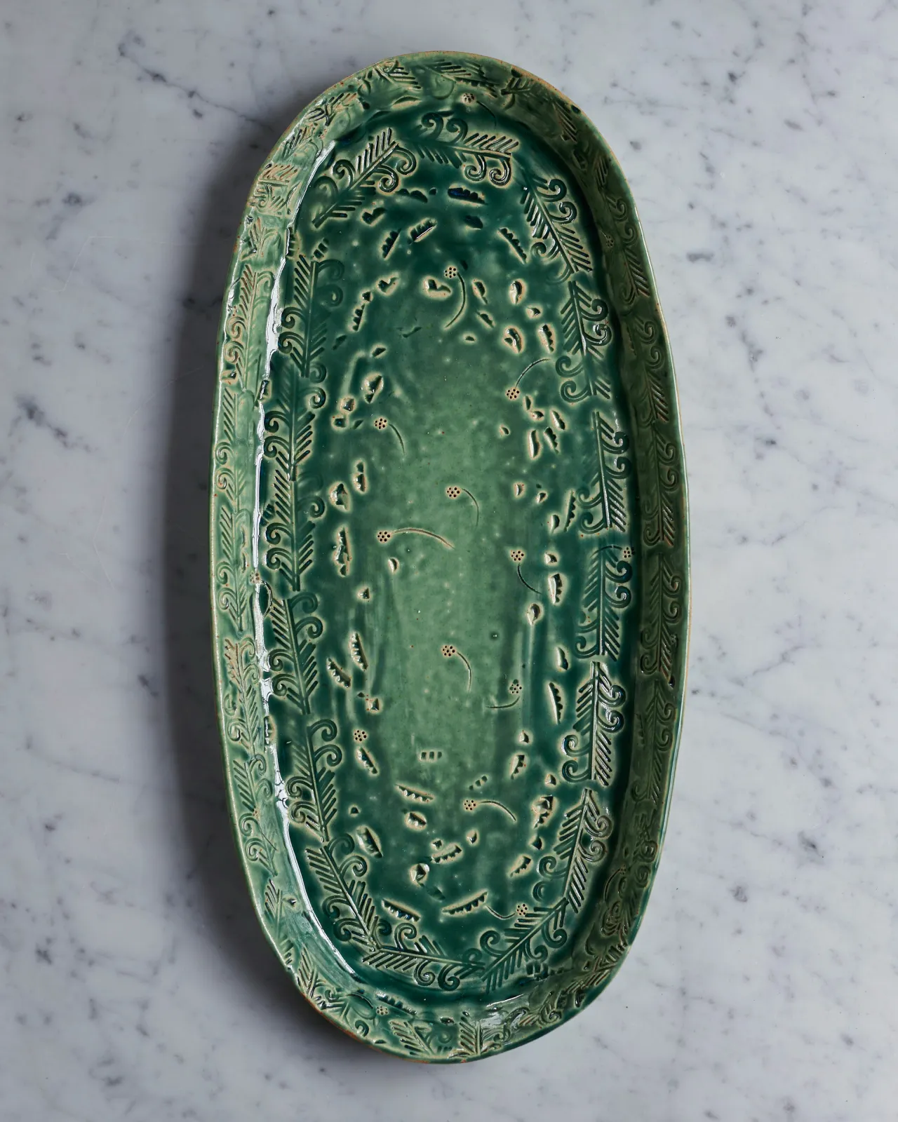 An overhead view of an oval-shaped handmade bohemian-style green ceramic platter with delicate impressions and surface patterns.