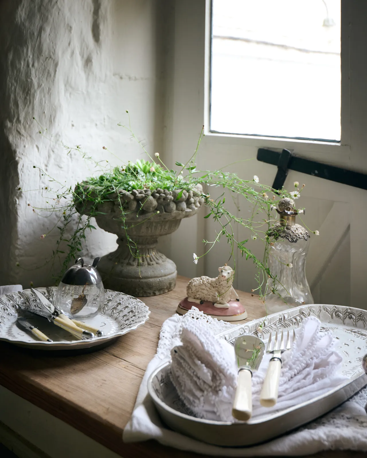 Two bohemian-style white ceramic platters, one oval-shaped, the other round, on a rustic farmhouse table with vintage silverware, white linens and wild succulent plant in a stone planter.