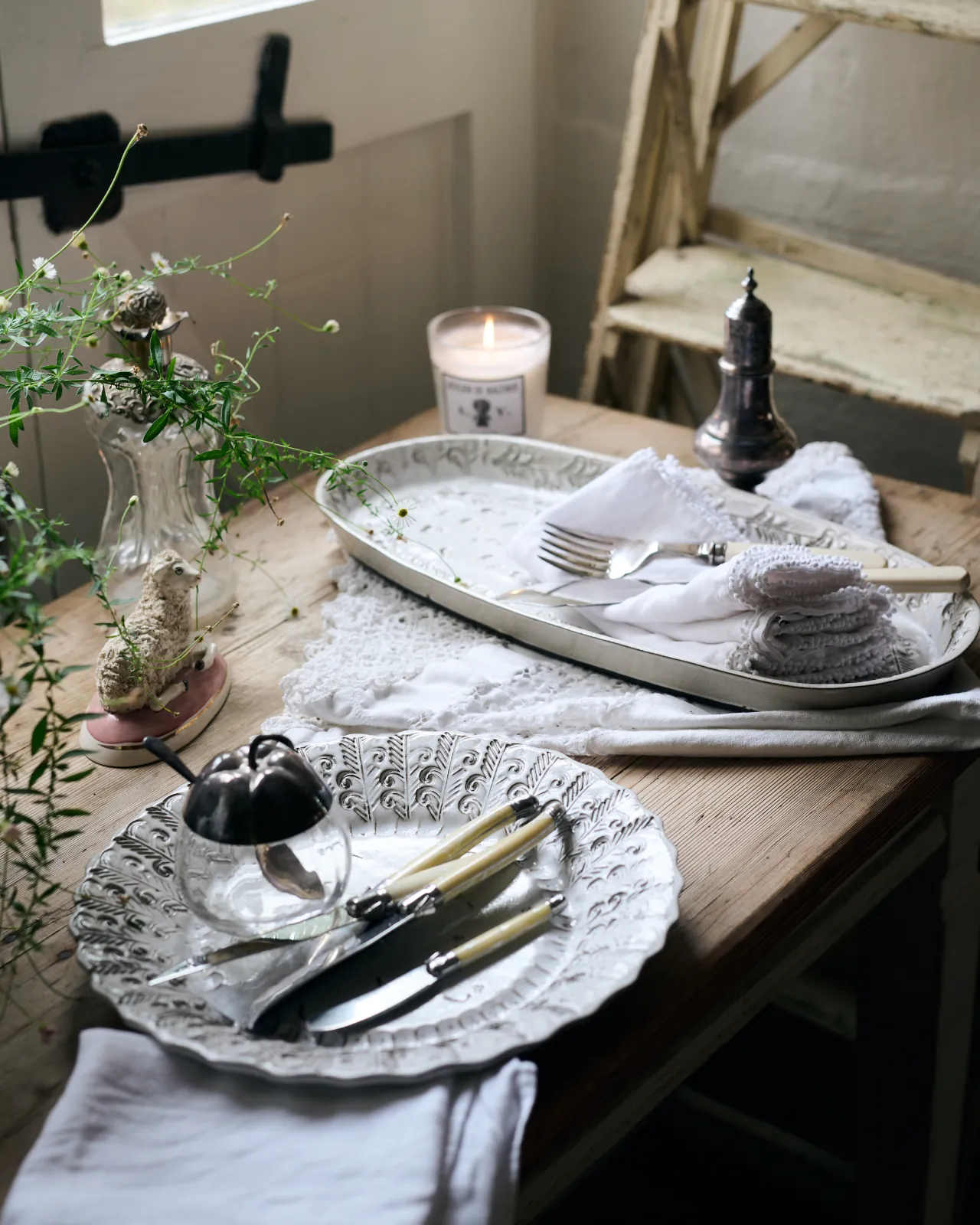 Two bohemian-style white ceramic platters, one oval-shaped, the other round, on a rustic farmhouse table with vintage silverware, white linens and a lit scented candle.