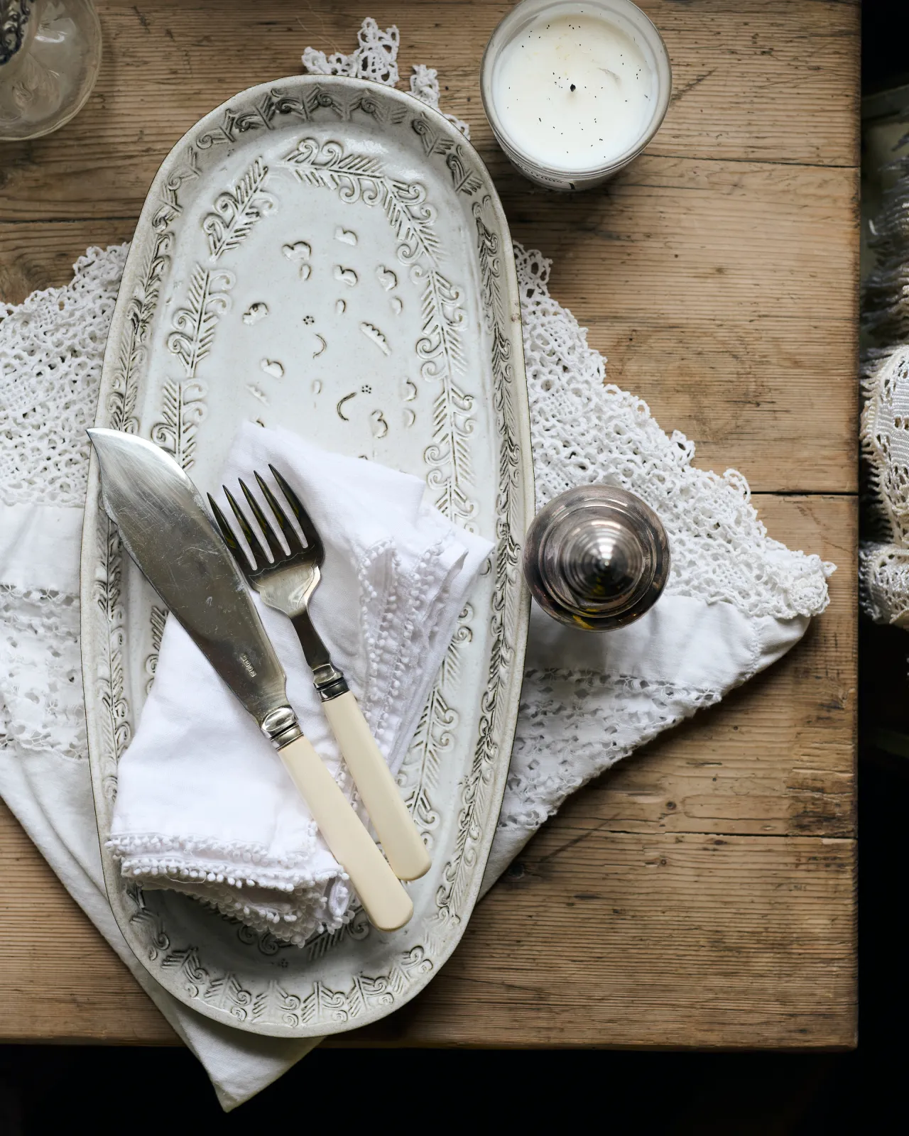 An overhead view of an oval-shaped handmade bohemian-style white ceramic platter with delicate impressions and surface patterns, complimented perfectly by vintage silverware, white linens and a rustic pine tabletop.
