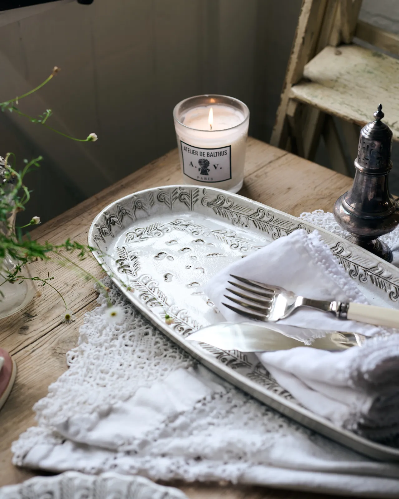 An oblique view of an oval-shaped handmade bohemian-style white ceramic platter with delicate impressions and surface patterns, complimented perfectly by vintage silverware, white linens and a rustic pine tabletop.