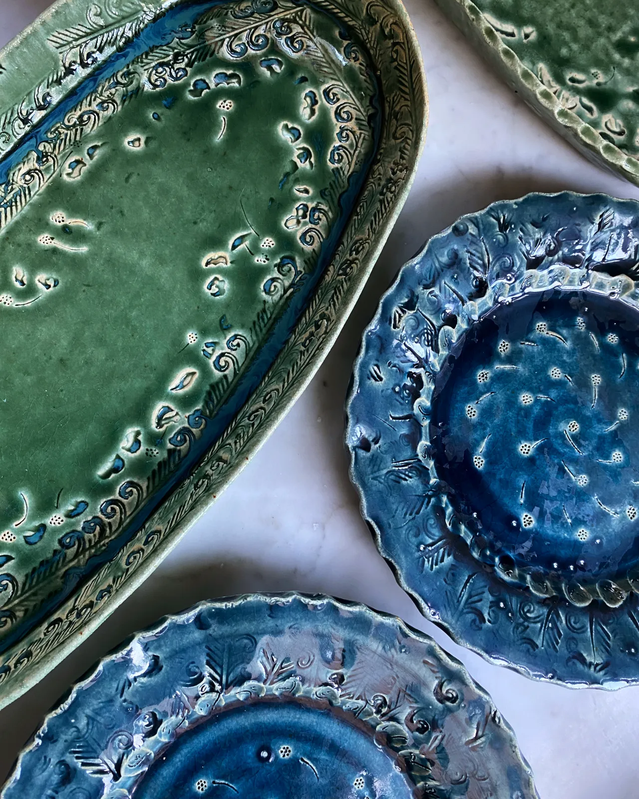 An overhead view of a green oval-shaped ceramic platter and two blue round-shaped ceramic platters, both with delicate impressions and surface patterns.