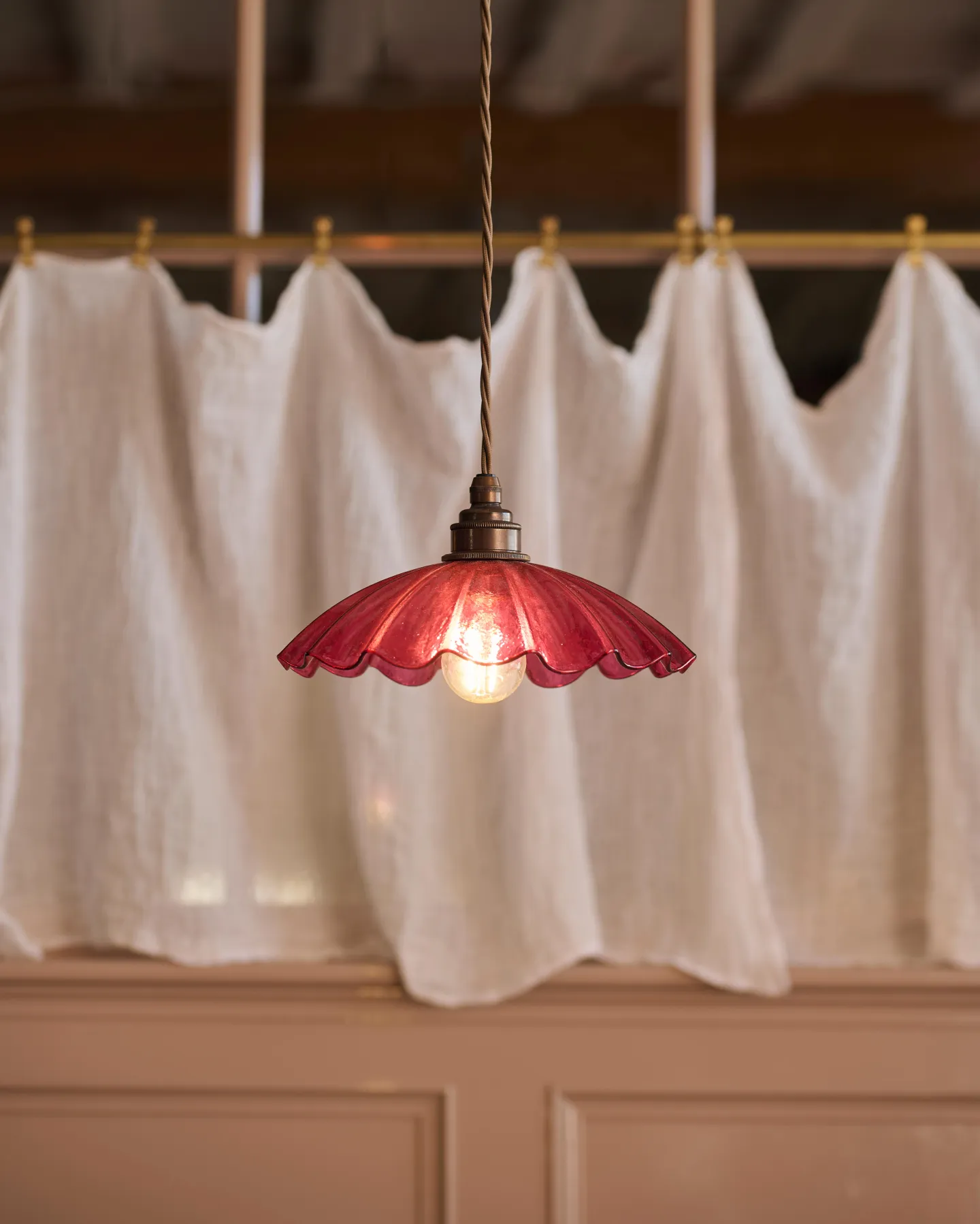 A small kitchen table with bench seating behind it with a pretty white curtain above it and a frilly cranberry pink, textured glass pendant light.