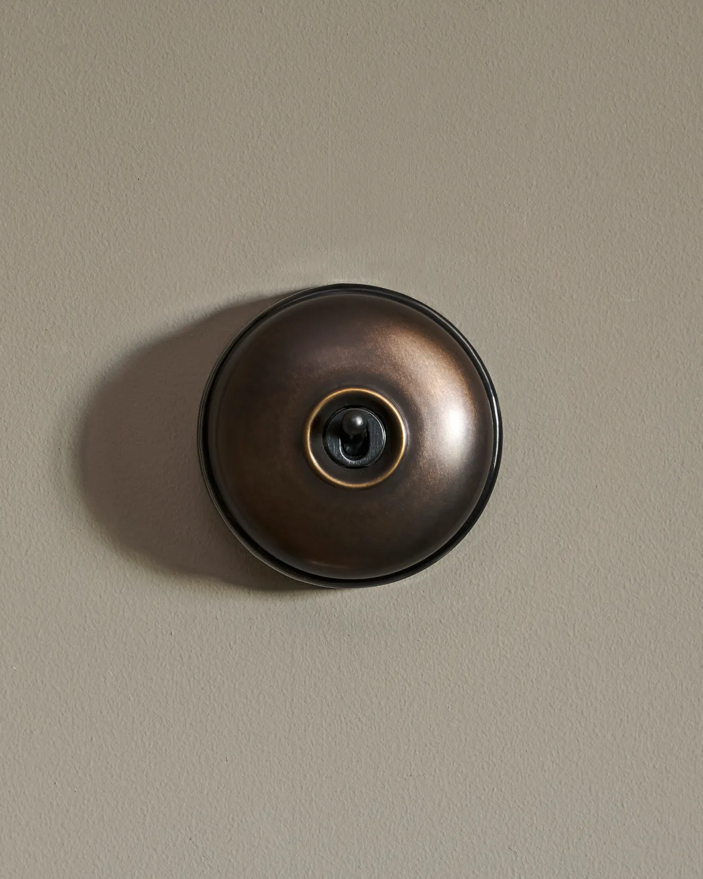 A straight-on view of a small 1 gang round-shaped toggle switch in a dark burnished brass finish with a black base.