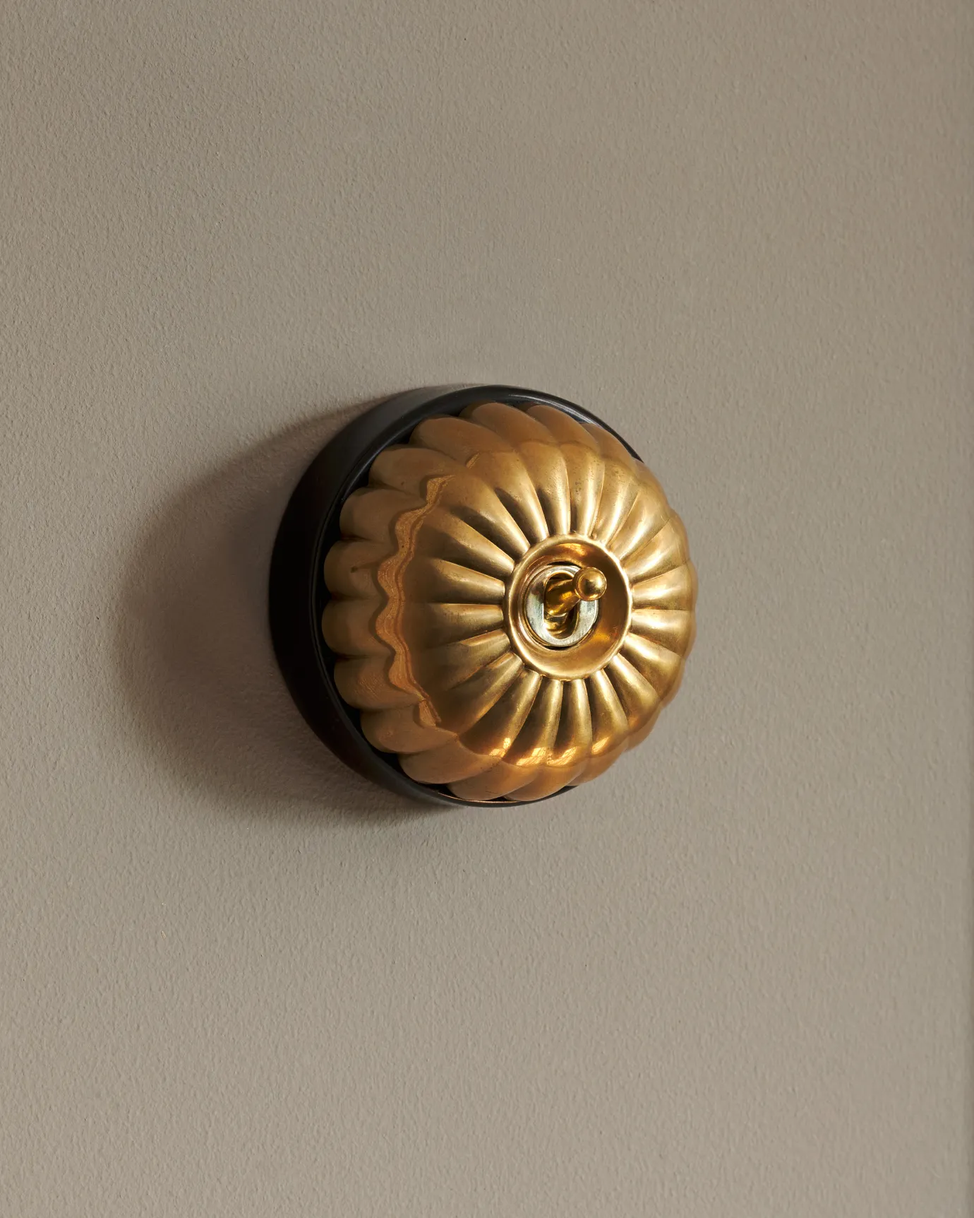 A side-on view of a small 1 gang round-shaped toggle switch in a warm shiny brass finish with a black base.