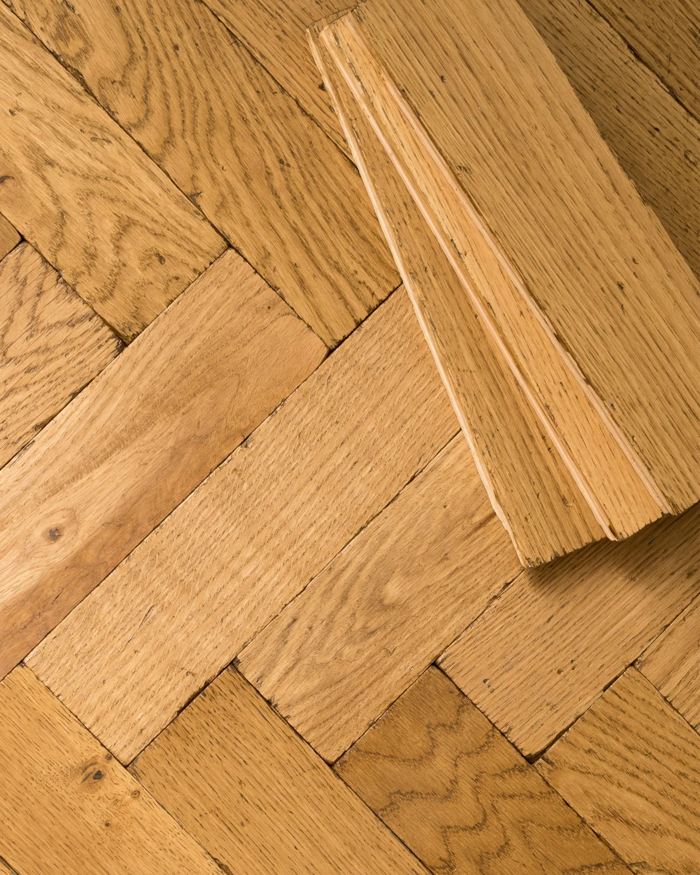 A close-up overhead view of oak parquet flooring with a warm aged finish. Three loose blocks are stacked in one corner.