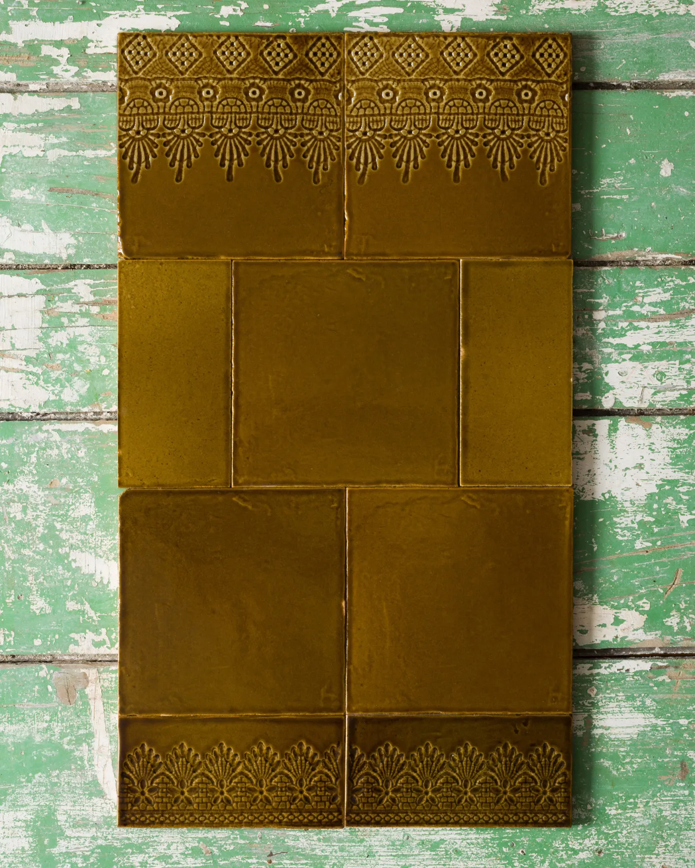 A small arrangement of handmade glossy sepia wall tiles - some plain, some with decorative lace patterns - on a rustic green and white tabletop.