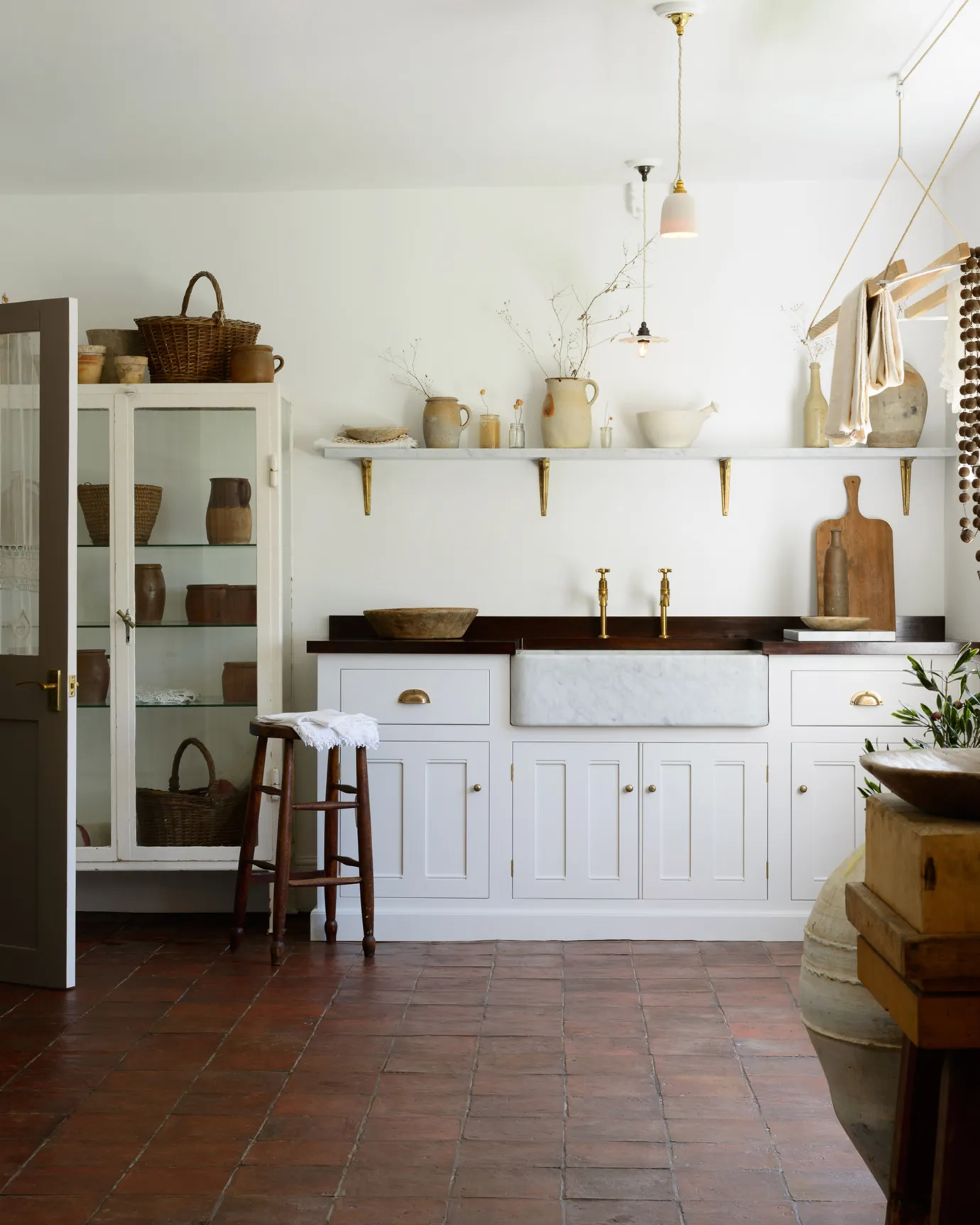 A Mediterranean-inspired scullery with white painted Georgian-style cabinetry, white walls, vintage furniture and square terracotta flooring with an authentic aged finish.