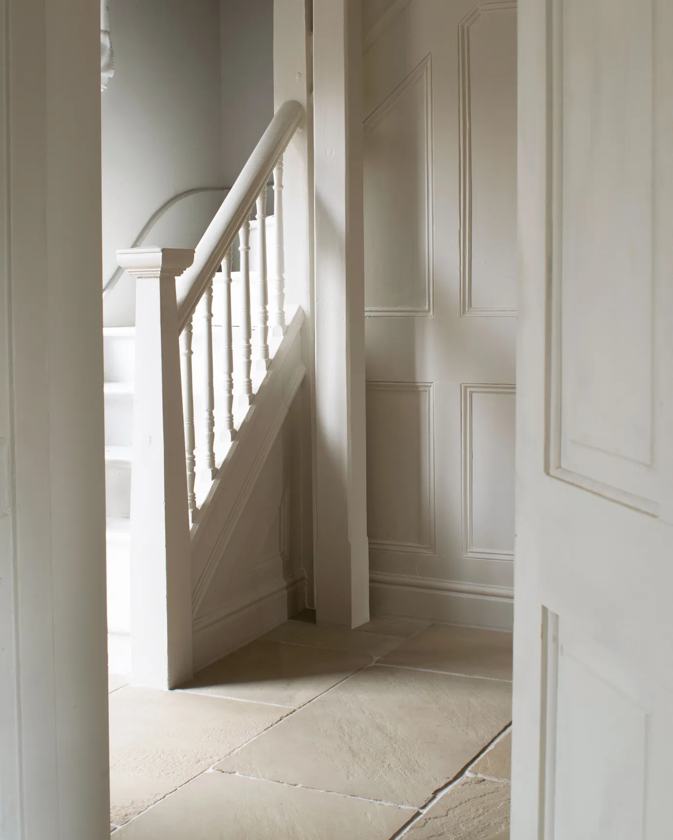 An oblique view of a classic stairway lobby with white walls, white painted woodwork and creamy coloured sandstone flooring.